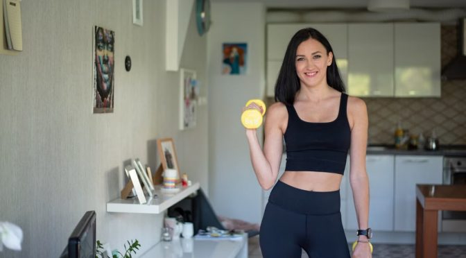 Smiling woman in activewear lifting dumbbells at home for a healthy lifestyle.